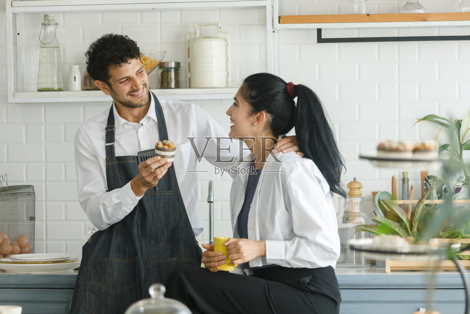 美丽、快乐、开朗的高加索夫妇在厨房系上围裙，带着爱的情感吃早餐。男女情人厨师。照片摄影图片