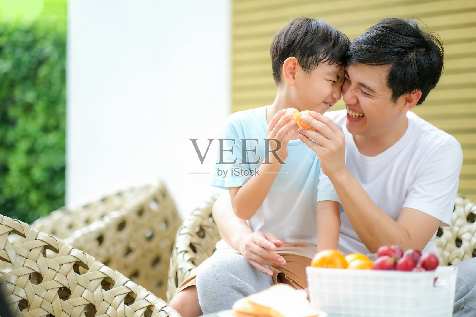 年轻的亚洲父亲碰碰橘子，头碰着爱、快乐和幸福的微笑，他们在花园里快乐地分享着饮料和食物。照片摄影图片