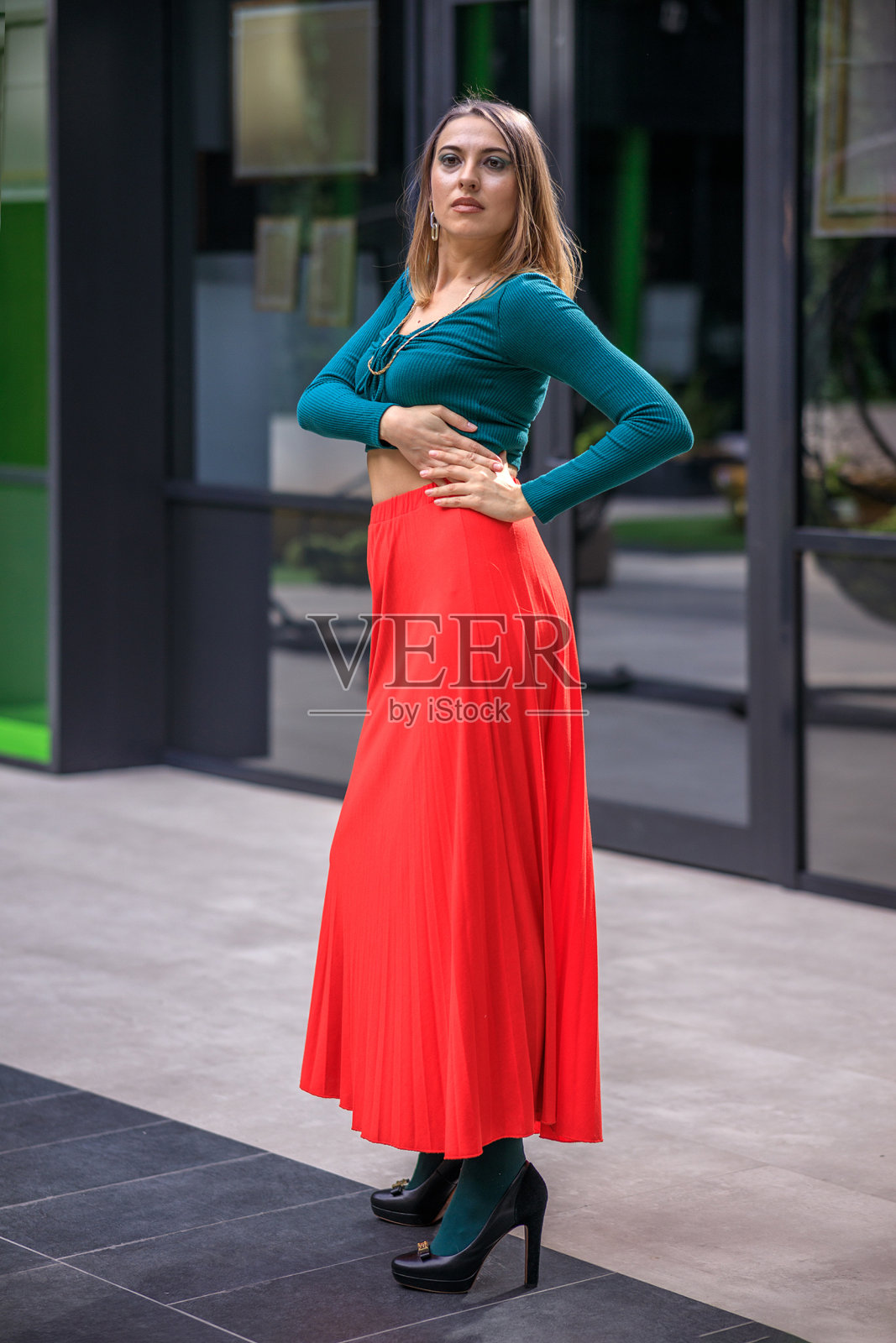 一个年轻的女人穿着鲜艳的衣服，红色的裙子和绿色的长袖站在一个商业中心。高跟鞋。照片摄影图片