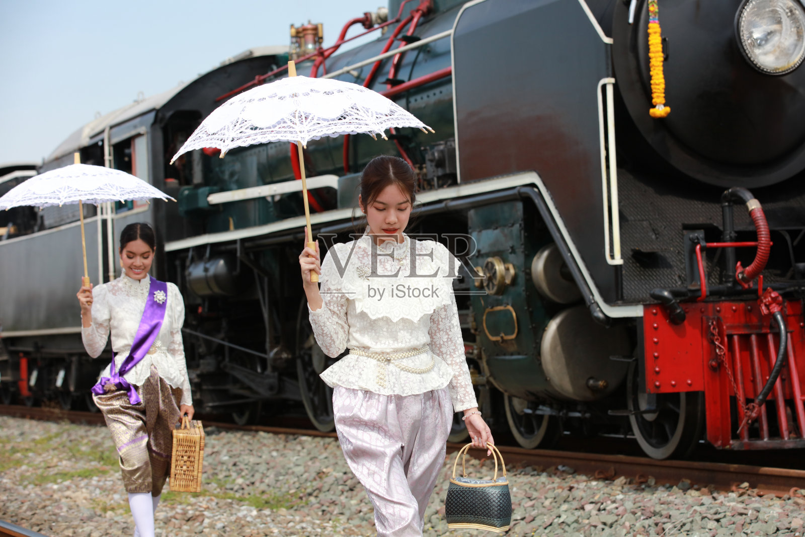 在泰国传统火车站，两名身穿泰国传统服装的亚洲妇女撑着雨伞走过蒸汽机车前的铁轨。照片摄影图片