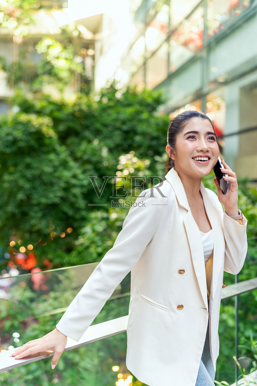 穿着白色西装的女性在现代城市背景下的城市绿化中打电话照片摄影图片