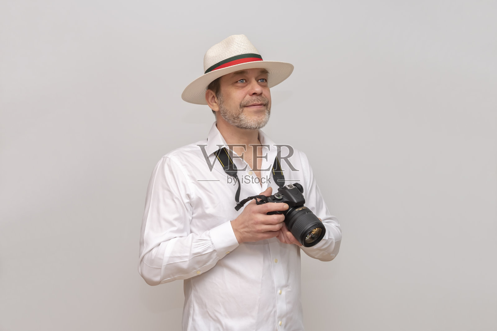 50-55岁的老人，戴着帽子，手里拿着相机，在白色的背景上看着远处。照片摄影图片