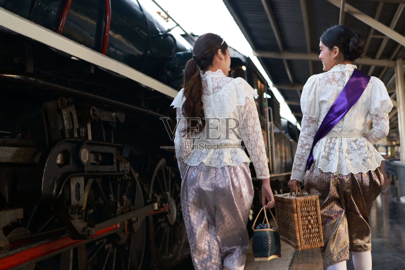 在复古火车站的月台上，两位穿着传统泰国服装的美女与蒸汽火车合影照片摄影图片