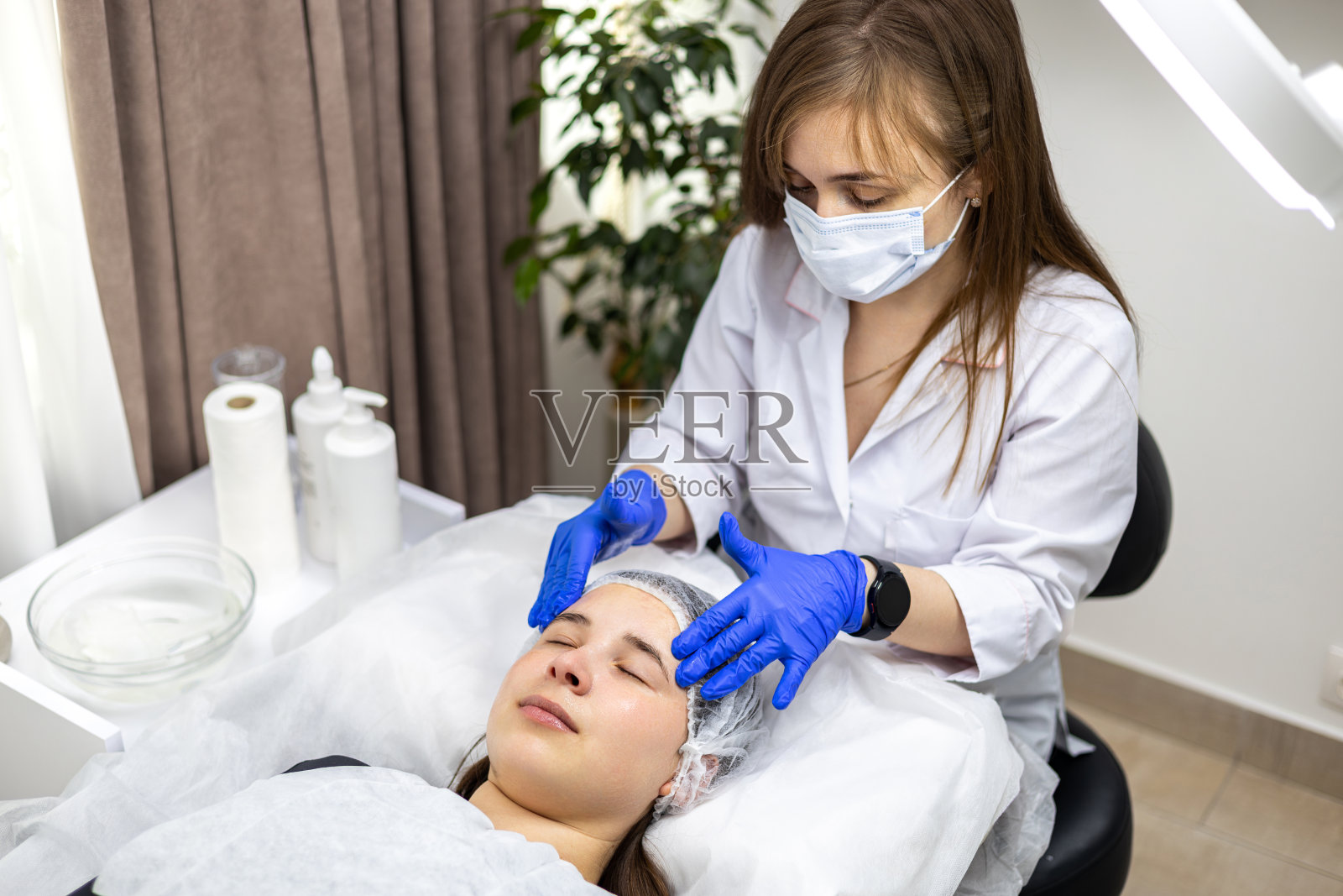 面部按摩是在美容院准备的。医生的手。治疗健康皮肤。一个年轻女人的头。清洗恢复活力。皮肤病面膜。排毒疗法照片摄影图片