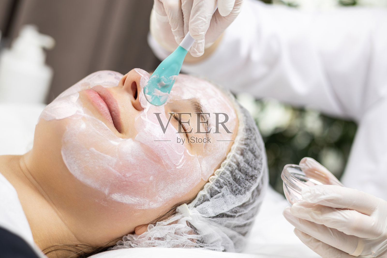 美丽的年轻女子微笑着放松着，而美容师正在用抹刀给她的脸上涂粘土面膜。照片摄影图片