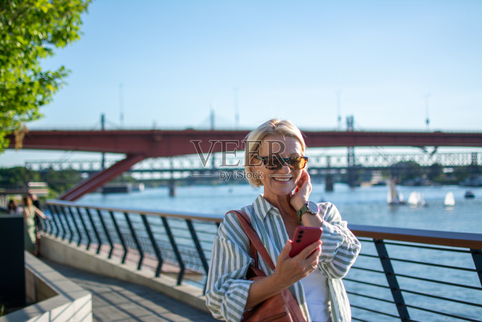 成熟的金发女人带着墨镜，微笑着在城市的河岸上享受照片摄影图片
