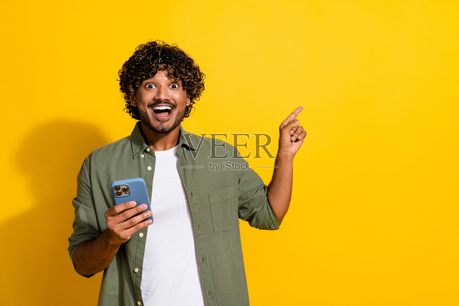 年轻有趣的卷发墨西哥人用他的智能手机的照片震惊浏览新闻直接手指网站孤立的黄色背景照片摄影图片