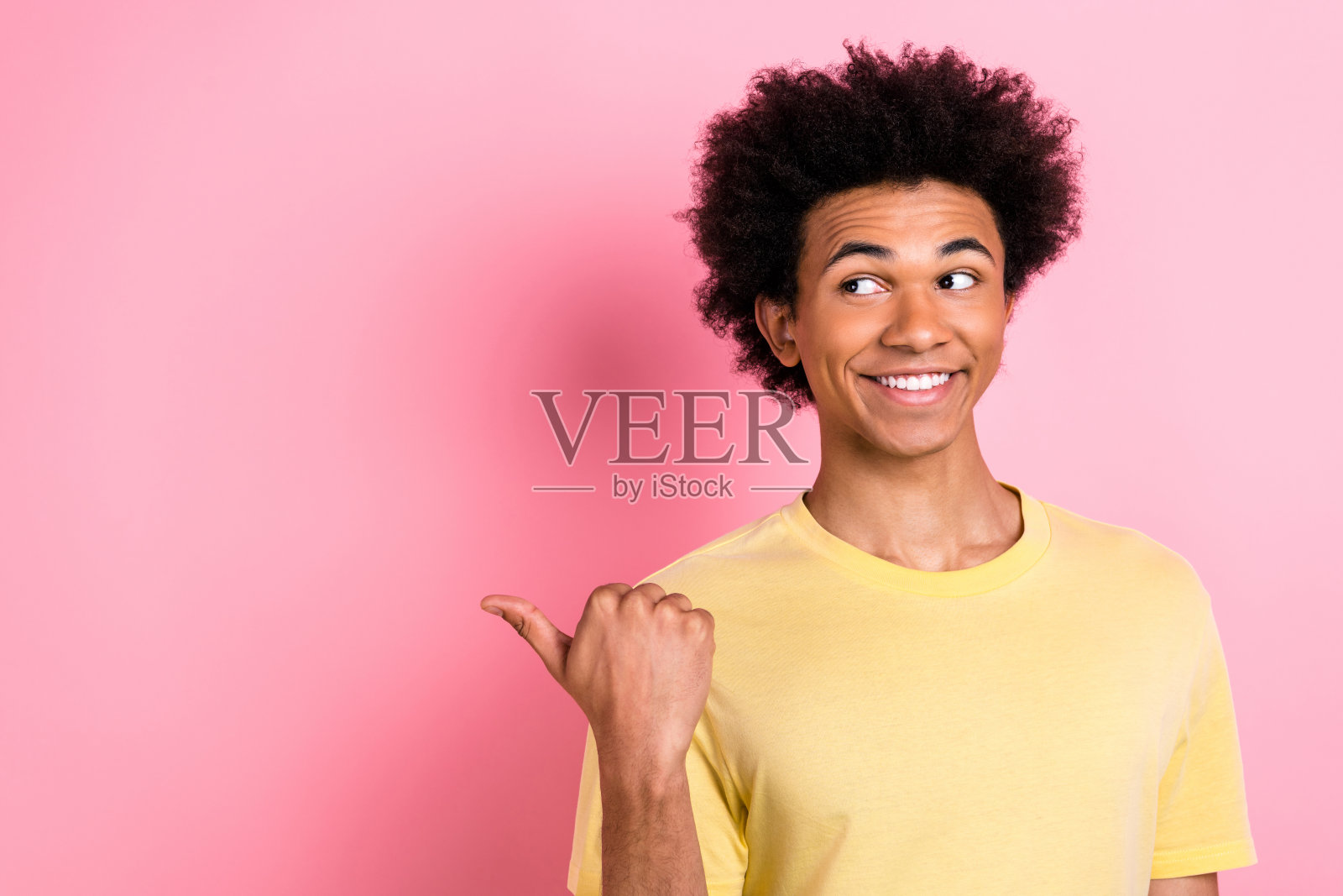 照片中时髦梦幻的男人穿着黄色t恤，手指着大拇指看着空旷的空间，孤立的粉红色背景照片摄影图片