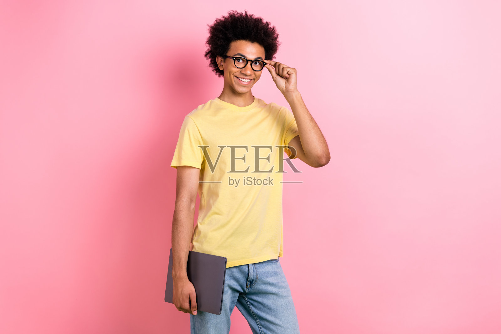 漂亮的年轻人拿着笔记本电脑，触摸眼镜，空着空间，穿着黄色t恤，孤立在粉红色的背景上照片摄影图片