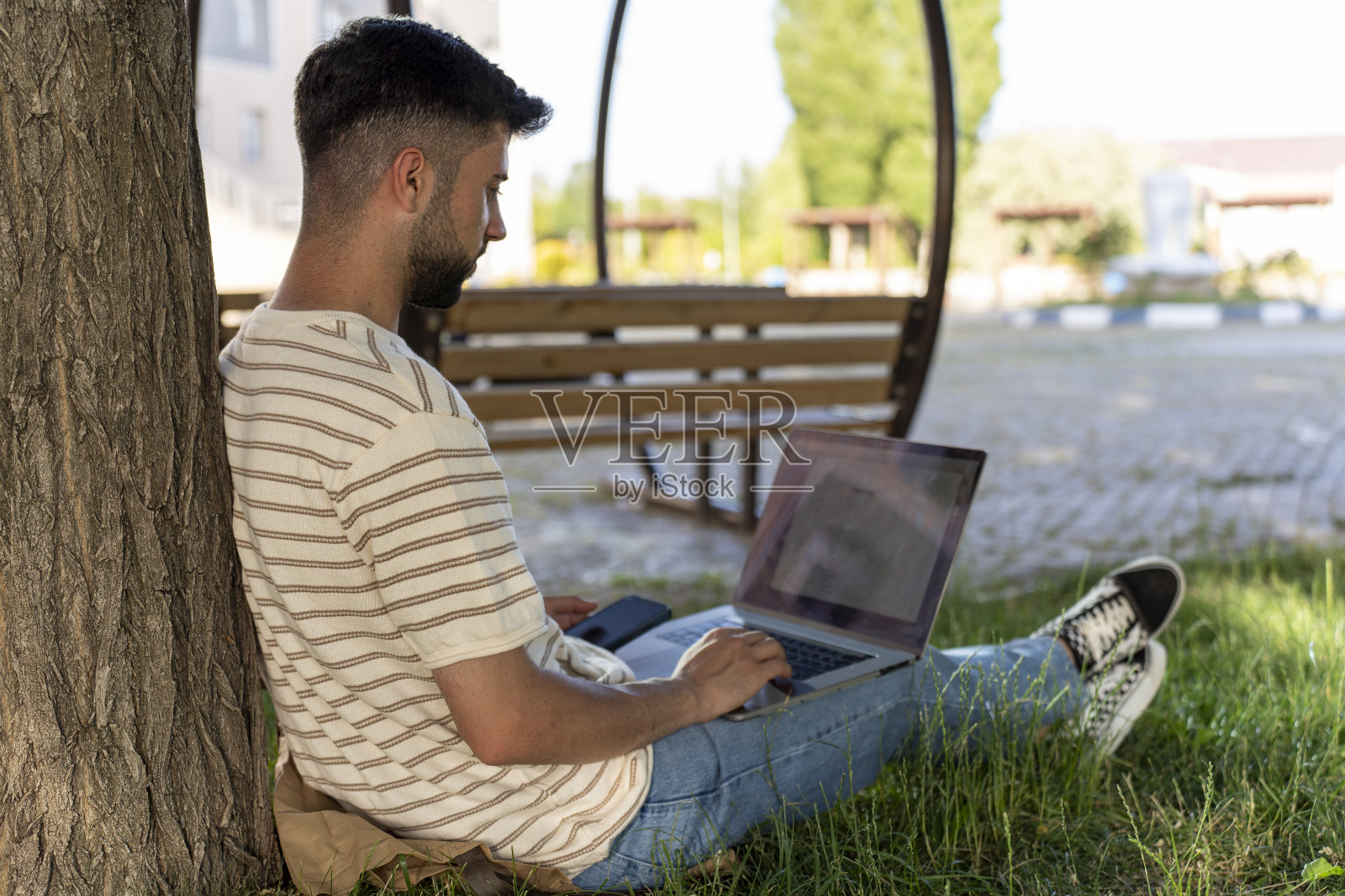 夏天的绿草坪上，一个年轻人拿着笔记本电脑坐在一棵树旁。照片摄影图片