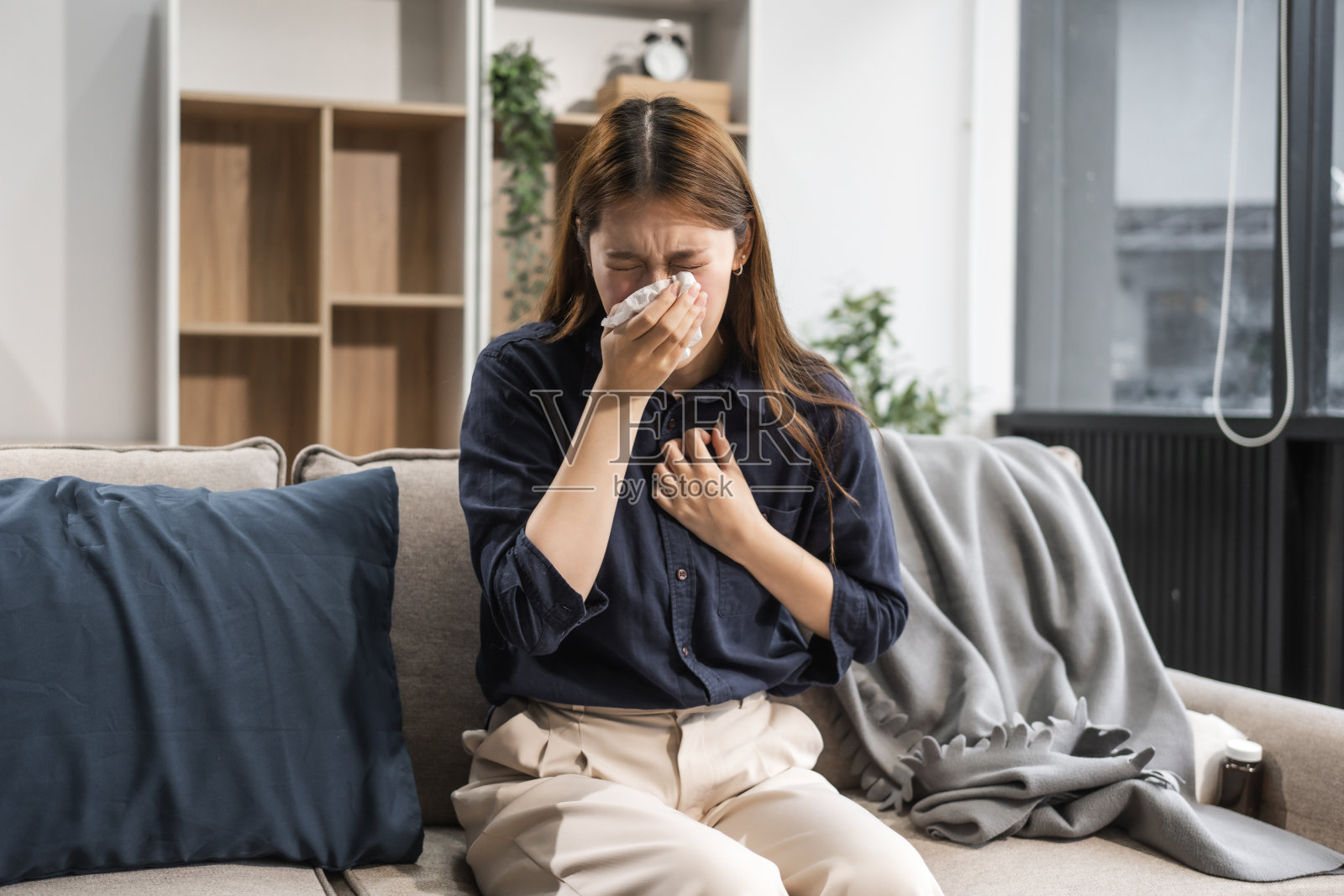 一位年轻的亚洲女性裹着围巾坐在沙发上。她有感冒症状，如咳嗽、打喷嚏、喉咙痛和充血，这表明她感染了病毒。照片摄影图片