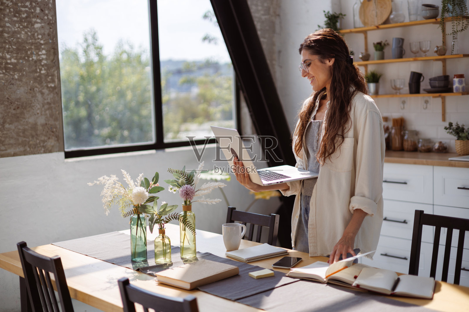 在家里的厨房里，微笑的女人拿着笔记本电脑，旁边放着书和咖啡照片摄影图片