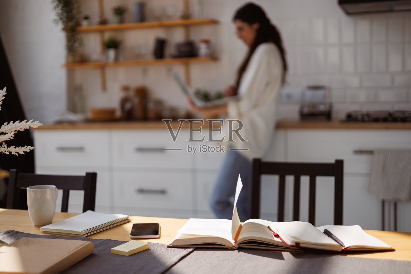 桌子上的智能手机和书，旁边的女人在家里的厨房里用笔记本电脑照片摄影图片