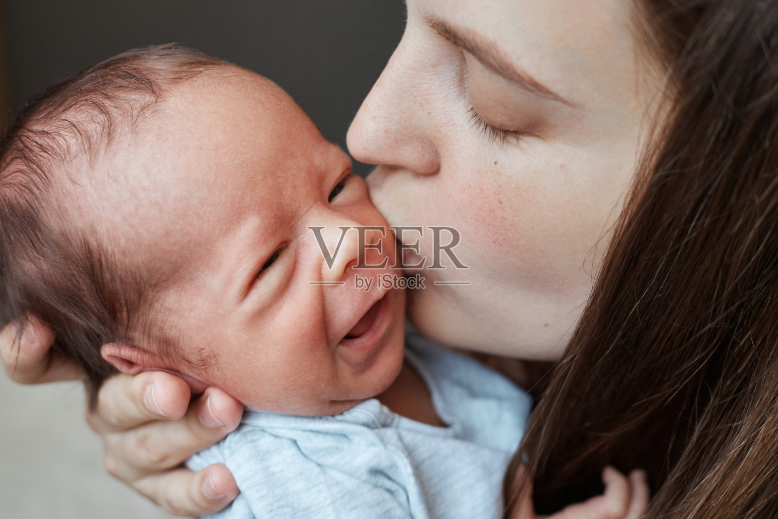 温柔的棕色头发的年轻白种人妈妈正在照顾她健康的新生儿，亲吻着她哭泣的小宝宝，宝宝安静地躺在妈妈的怀里照片摄影图片