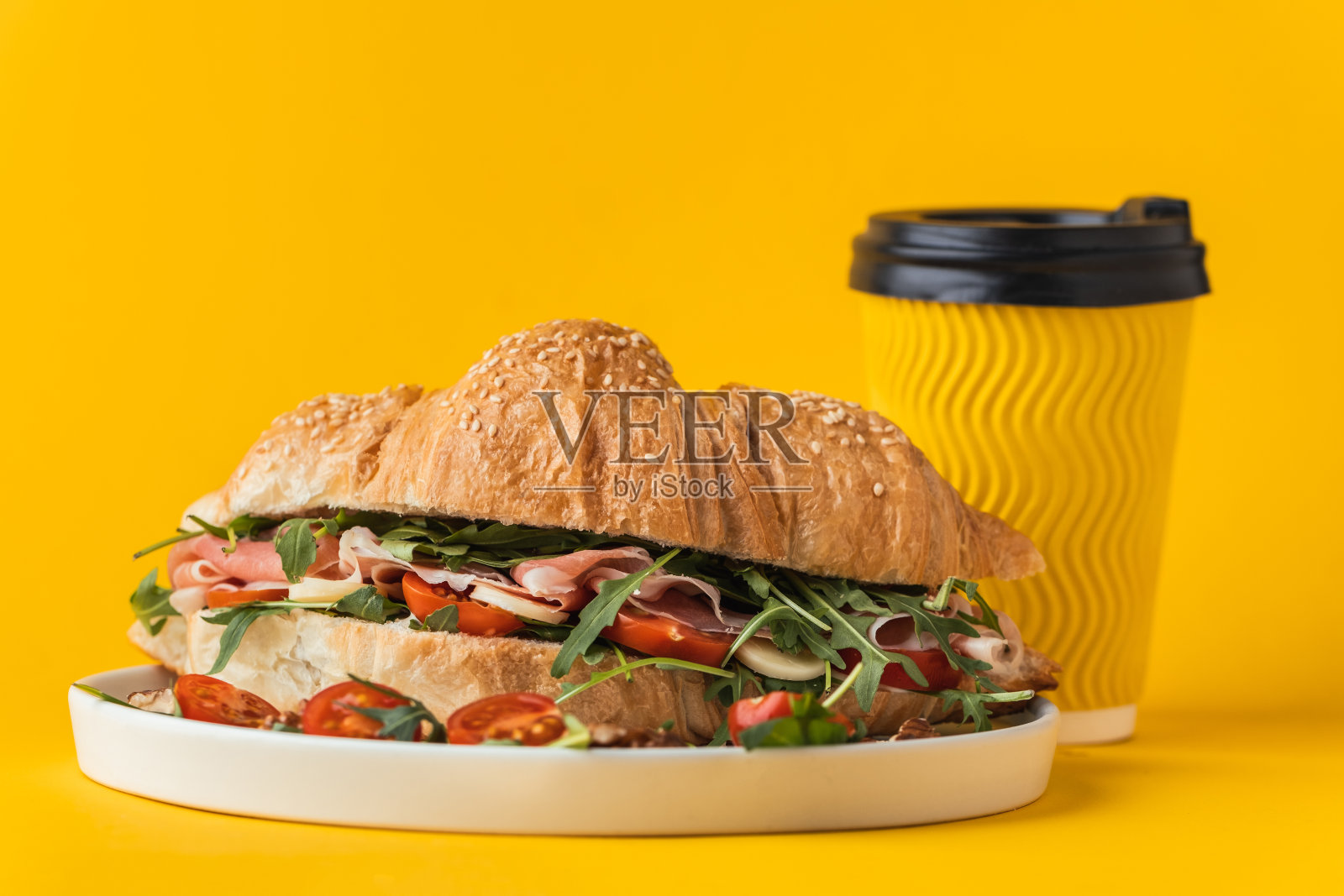 新鲜出炉的牛角面包配伊比利亚火腿、西红柿和沙拉馅，黄色背景。牛角面包的模拟旁边的黄色纸杯咖啡与复制空间的免费文本照片摄影图片
