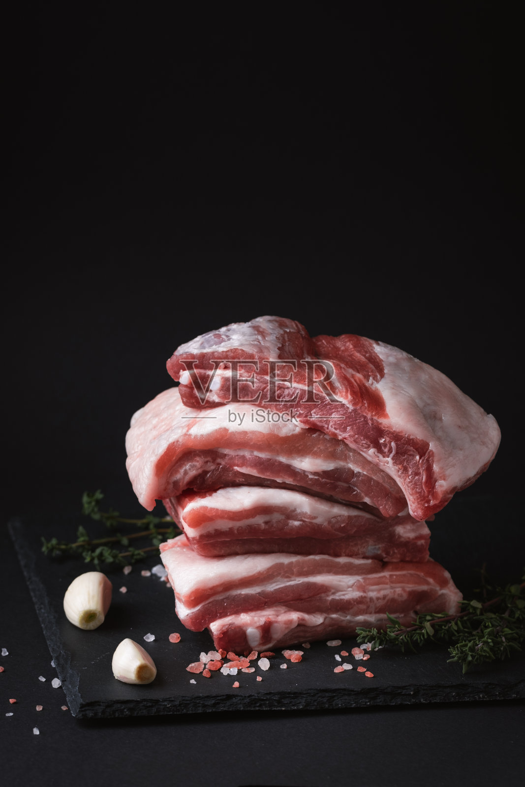 近距离拍摄的生猪肉，周围的盐晶体，大蒜和香料在黑色的背景。为自由文本复制空间。烹饪肉类菜肴的食谱模板。照片摄影图片