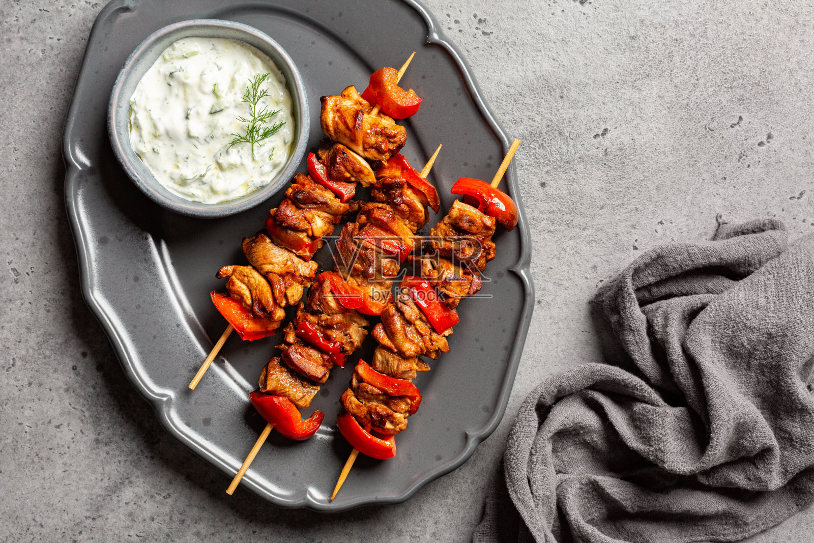 地中海烤鸡串的俯视图，由腌制的鸡肉和红灯笼椒制成。由酸奶、黄瓜碎、柠檬汁、橄榄油制成的希腊酸奶酱。灰色的桌子表面和餐巾。照片摄影图片