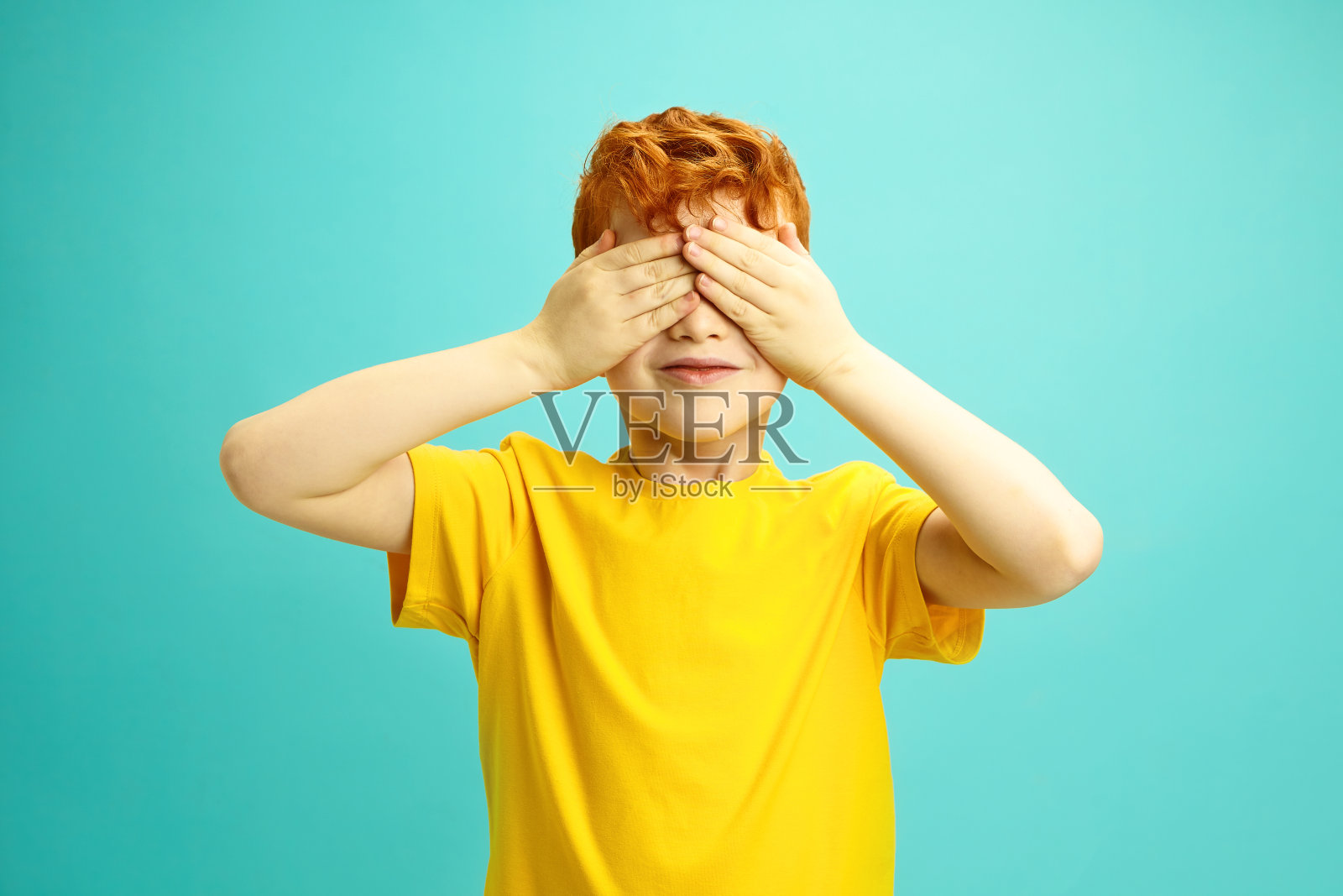 爱尔兰裔小男孩的工作室肖像，卷曲的红色头发用两只手遮住眼睛，穿着黄色t恤，站在蓝色孤立的背景上。照片摄影图片