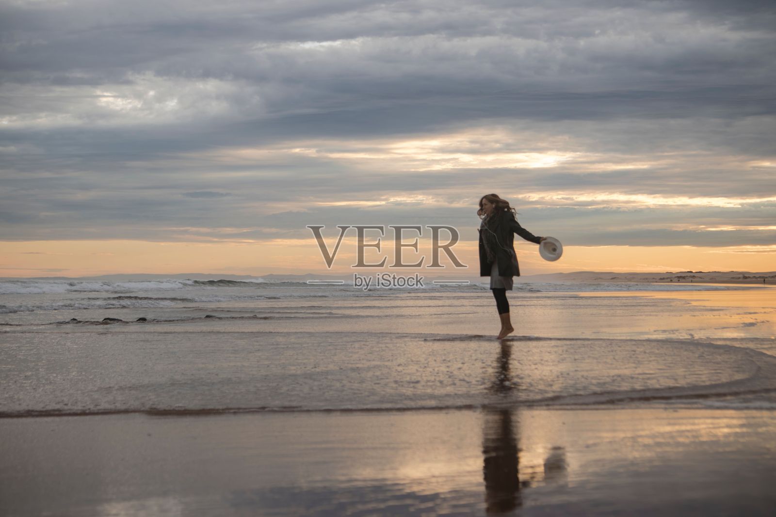 黄昏时分，多云的天空中，一名女子拿着帽子在海滩上跳跃照片摄影图片