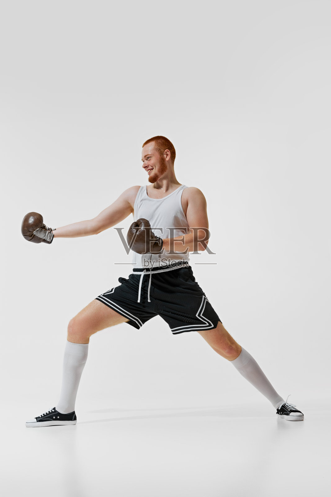 在白色的背景上，一个穿着休闲服、面带微笑、竞技的年轻人，戴着老式的棕色拳击手套在运动，孤立地训练着。照片摄影图片