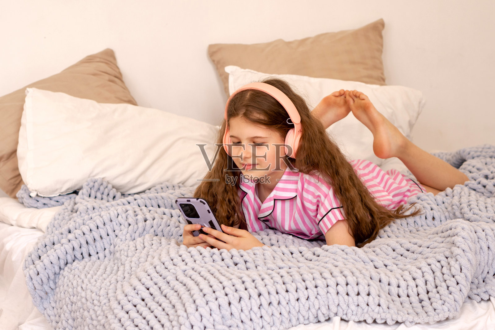 穿着粉色睡衣的女孩躺在床上玩智能手机。照片摄影图片