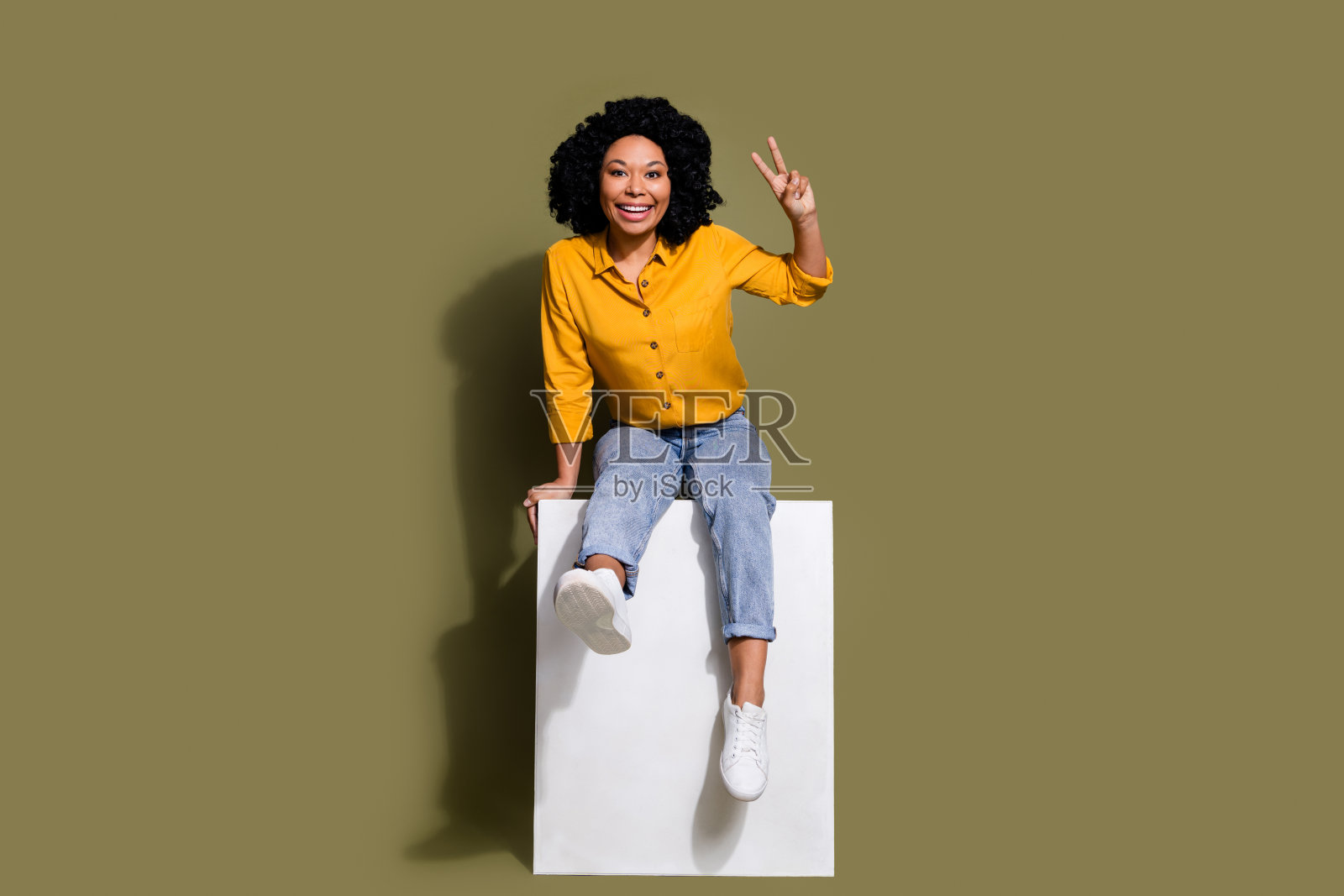 漂亮女士的全身肖像坐在立方体显示v字穿黄色衬衫孤立的卡其色背景照片摄影图片