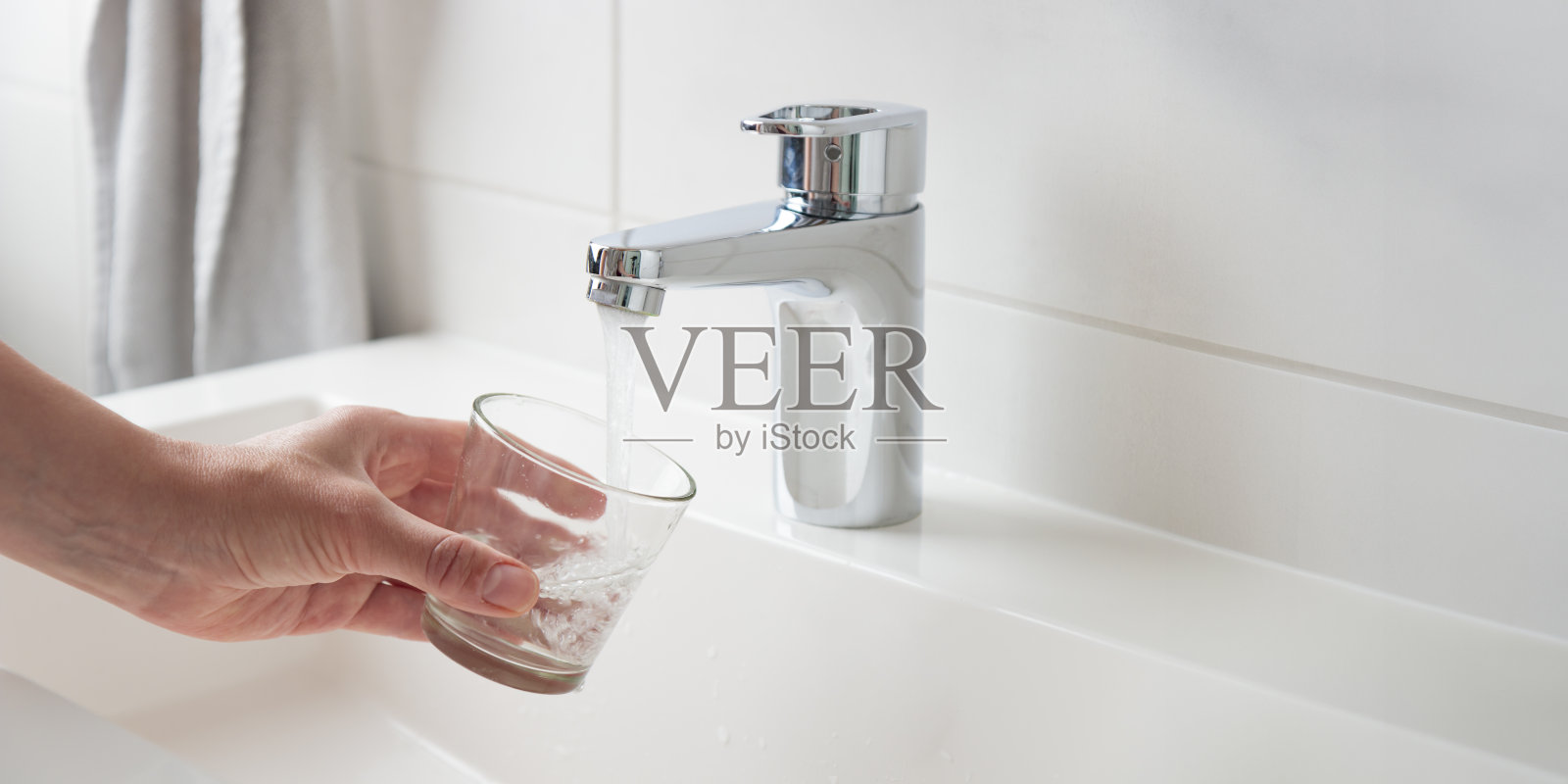在白色的浴室里，口渴的女人从水龙头往玻璃杯里倒入新鲜纯净的可饮用的自来水，在家里打着自然洁净水的概念照片摄影图片