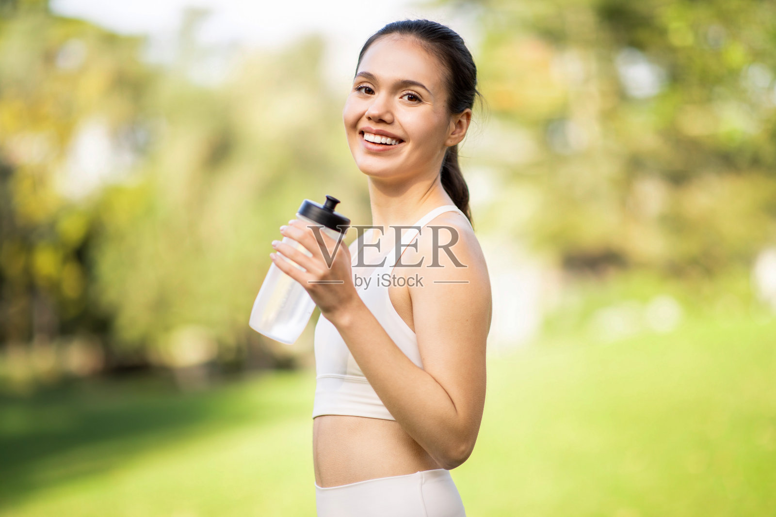 一个扎着马尾辫、容光焕发的年轻女子，穿着白色健身服，手里拿着一个水瓶，笑容灿烂照片摄影图片