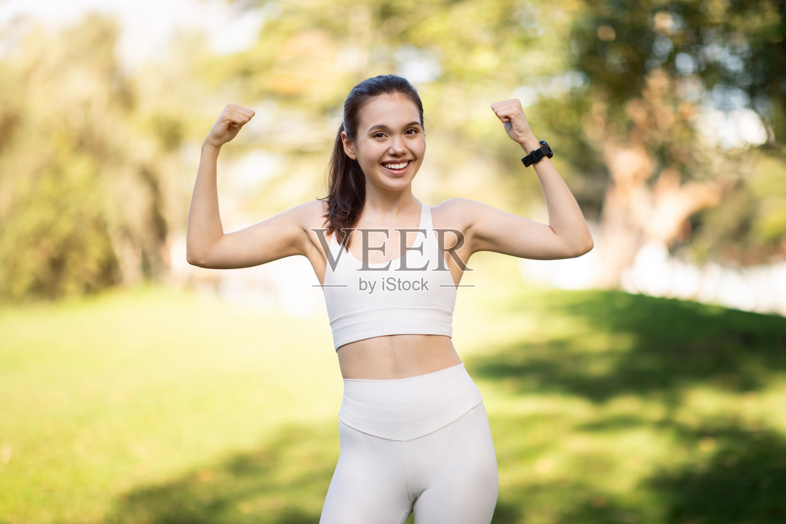 一个快乐的年轻女子穿着白色运动胸罩和打底裤在阳光明媚的公园里展示她的肌肉照片摄影图片