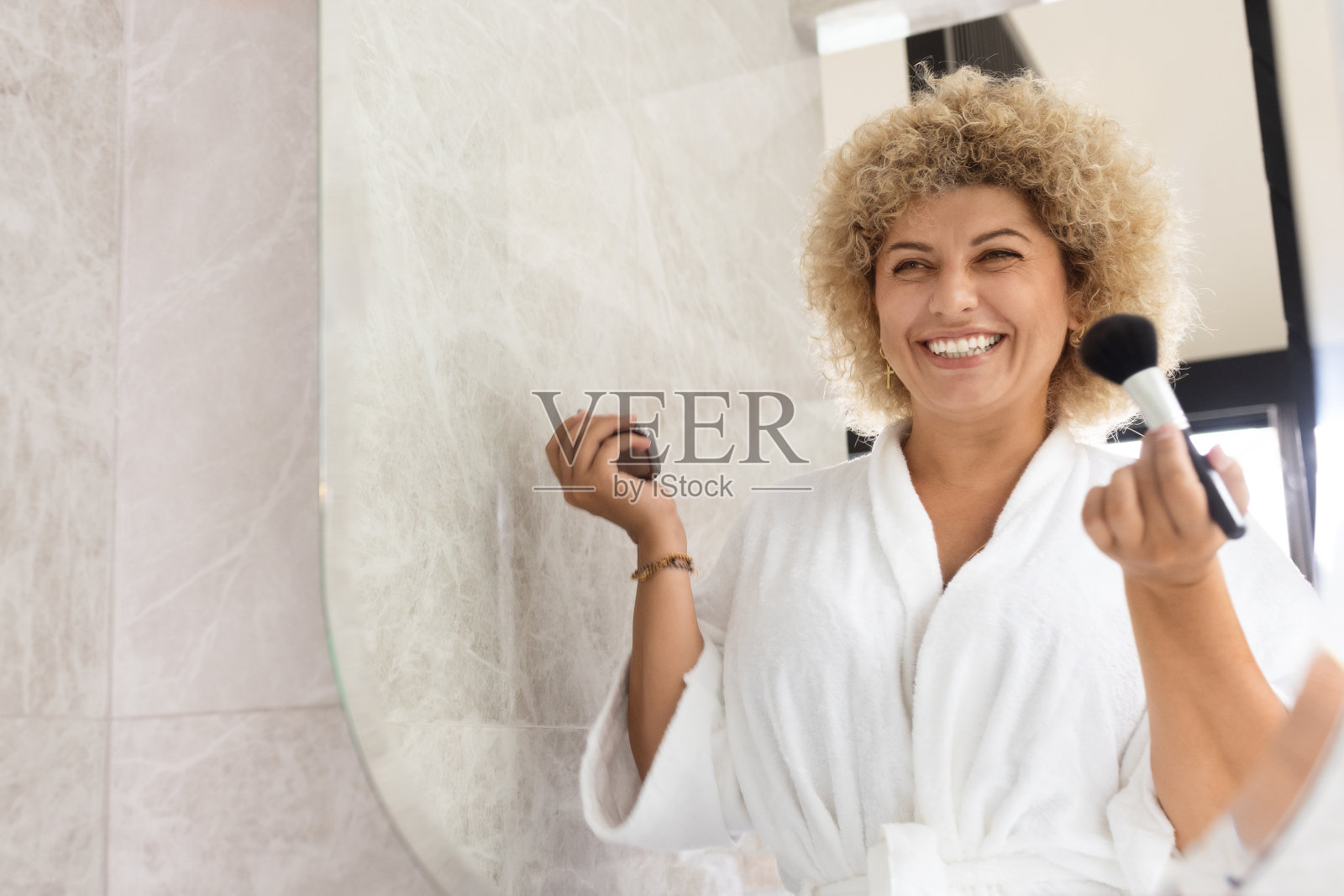 一位穿着白色浴袍的开朗的年轻女子在光线充足的浴室里化妆，享受着早晨的美容程序，散发着积极的光芒。照片摄影图片