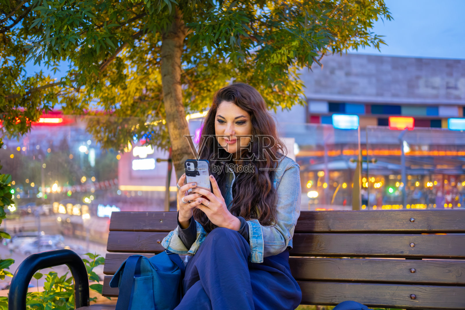 微笑的女人在城市享受夜晚，在公园的长椅上使用智能手机照片摄影图片