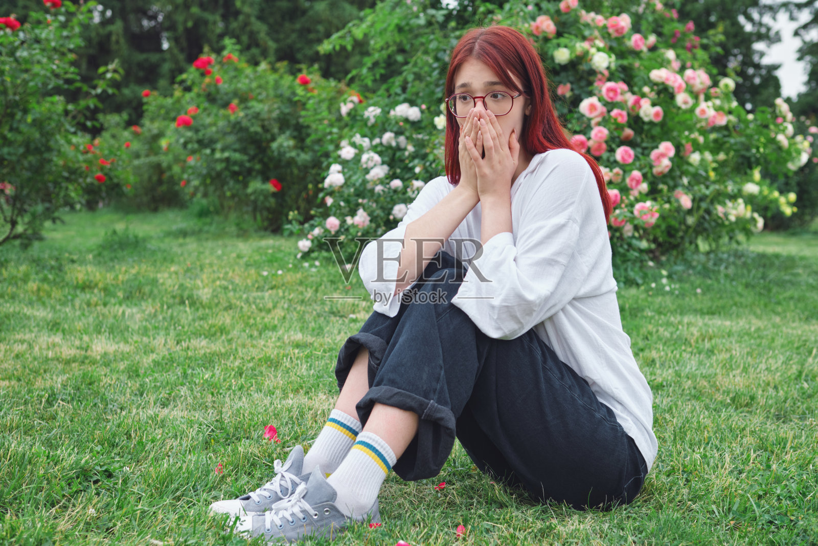 一个红头发戴眼镜的十几岁女孩坐在盛开的花园里，用手捂住鼻子，对难闻的气味或过敏做出反应照片摄影图片