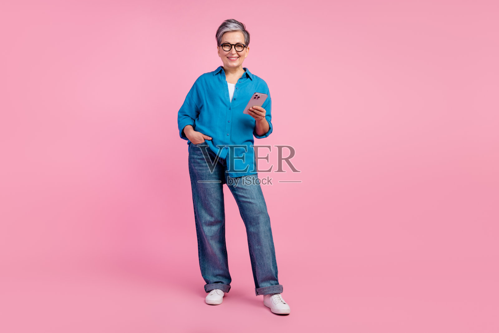 迷人的老年妇女的全身照片拿着电话，穿着时髦的蓝色衣服，孤立在粉红色的背景上照片摄影图片