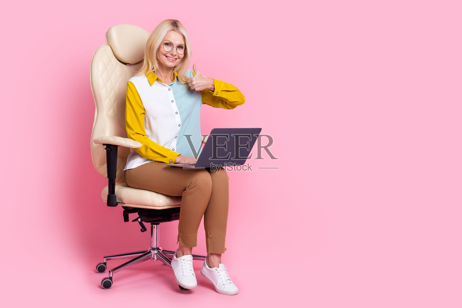 漂亮老太太的全身肖像，坐在椅子上网本大拇指朝上，穿着衬衫孤立在粉红色的背景照片摄影图片