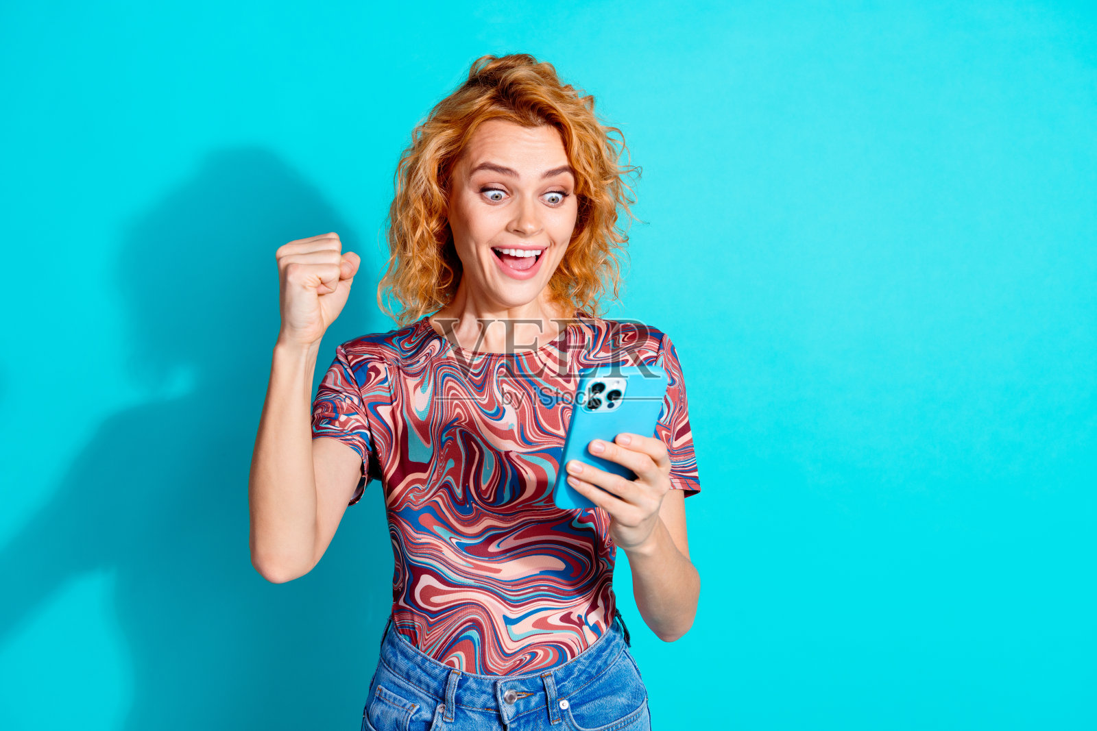 漂亮女士的肖像，用智能手机举起拳头，穿着t恤，孤立在蓝绿色的背景上照片摄影图片