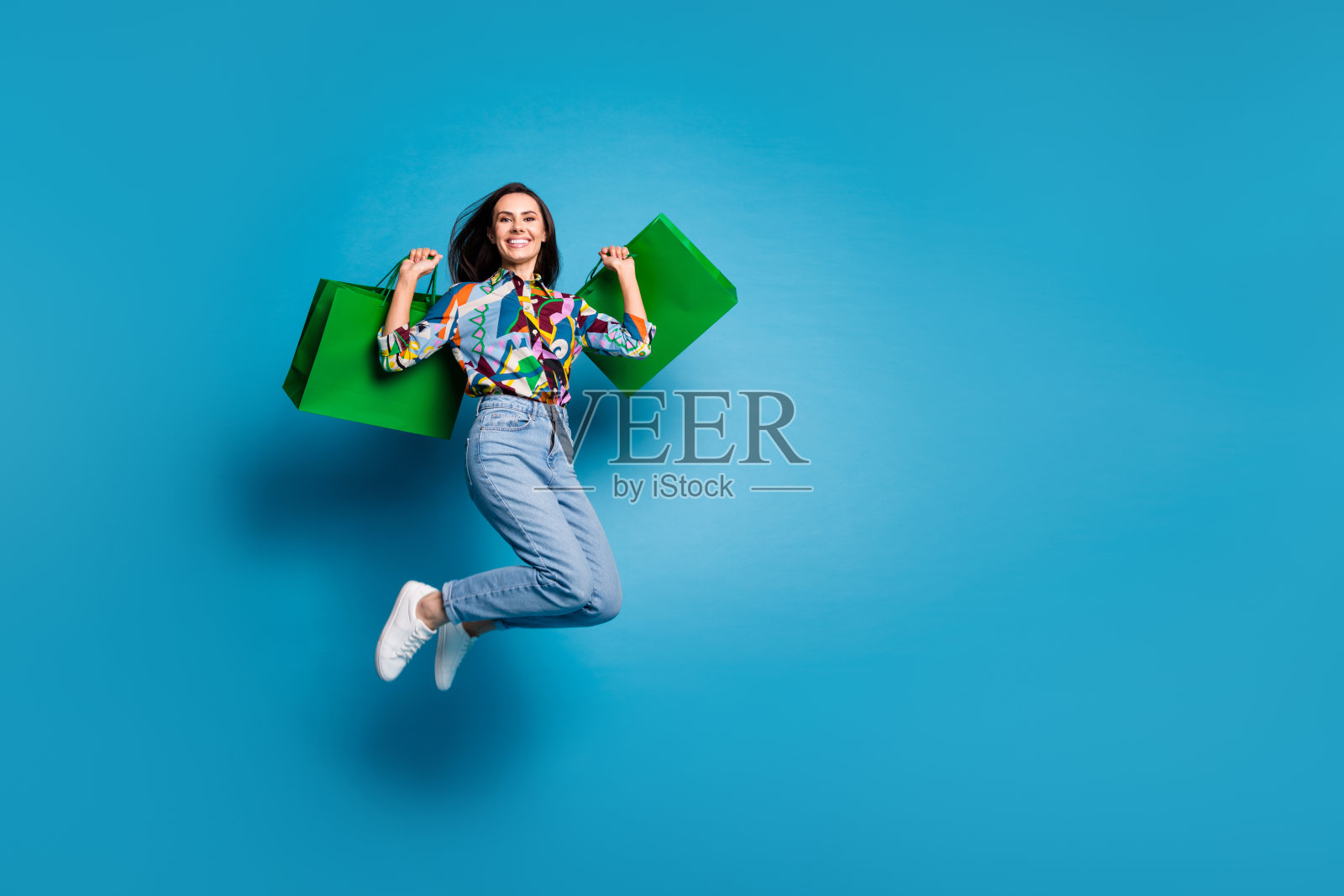全长照片正面华丽可爱的女人穿印花衬衫牛仔裤抱着购物袋跳跃孤立的蓝色背景照片摄影图片