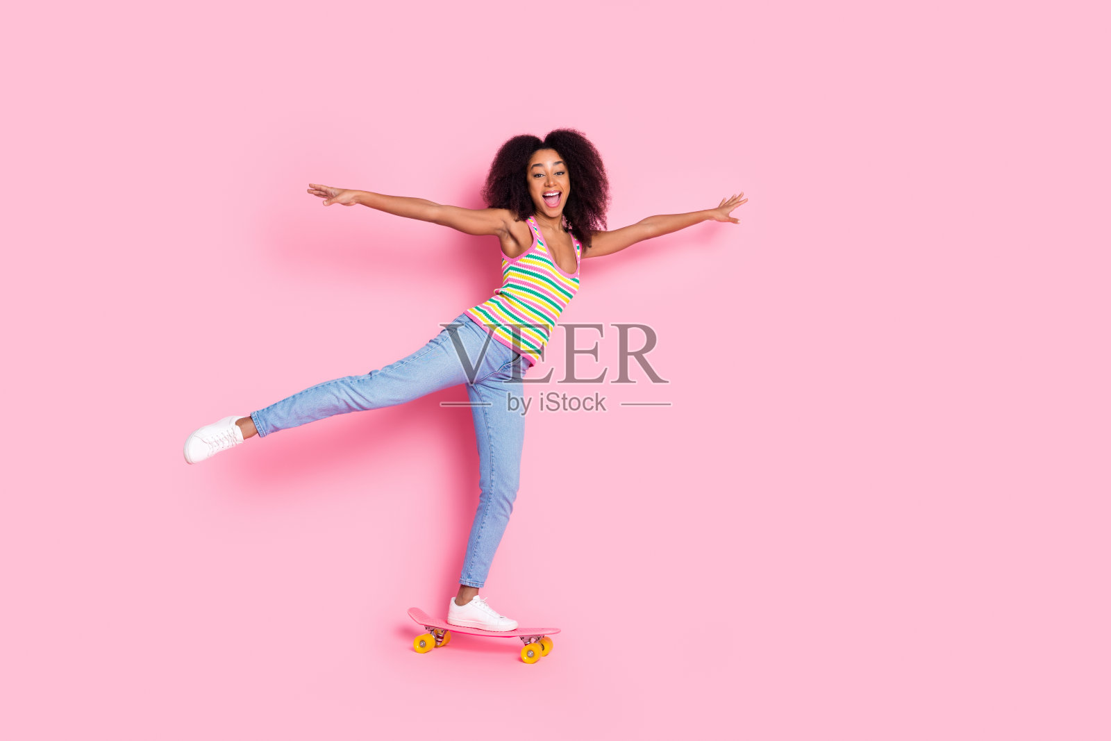 可爱可爱的卷发女孩的全长照片穿彩色顶骑滑板有乐趣孤立在粉红色的背景照片摄影图片