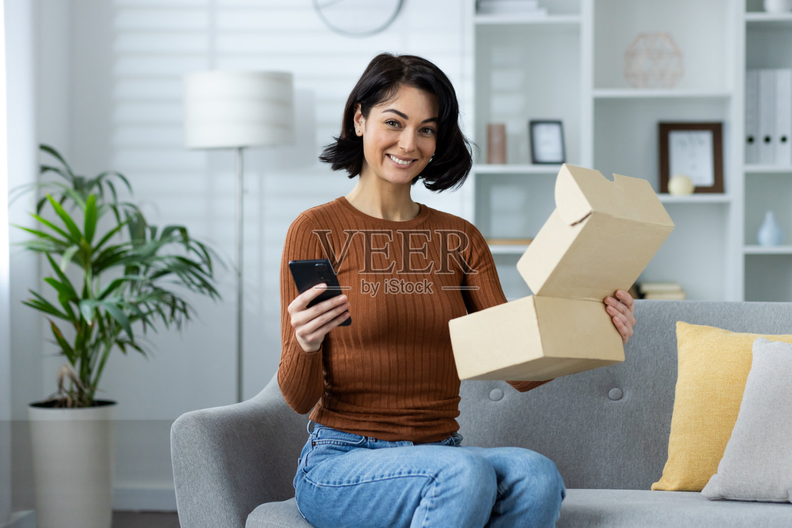 一个微笑的年轻女子的肖像，穿着休闲的衣服在家里，坐在沙发上，拿着一个电话和一个包裹，一个打开的盒子，看着相机照片摄影图片