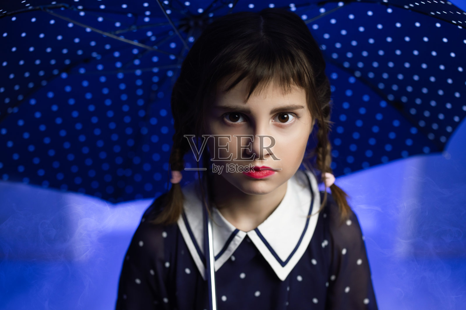 一个女孩梳着哥特式的辫子，手里拿着一把伞，背景是蓝色的。穿着裙子扎着小辫的女孩看着镜头。紫色背景和烟雾照片摄影图片