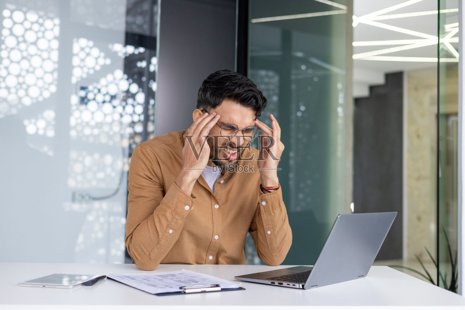 疲惫的年轻印度男子在办公室伏案工作，双手抱头缓解压力照片摄影图片