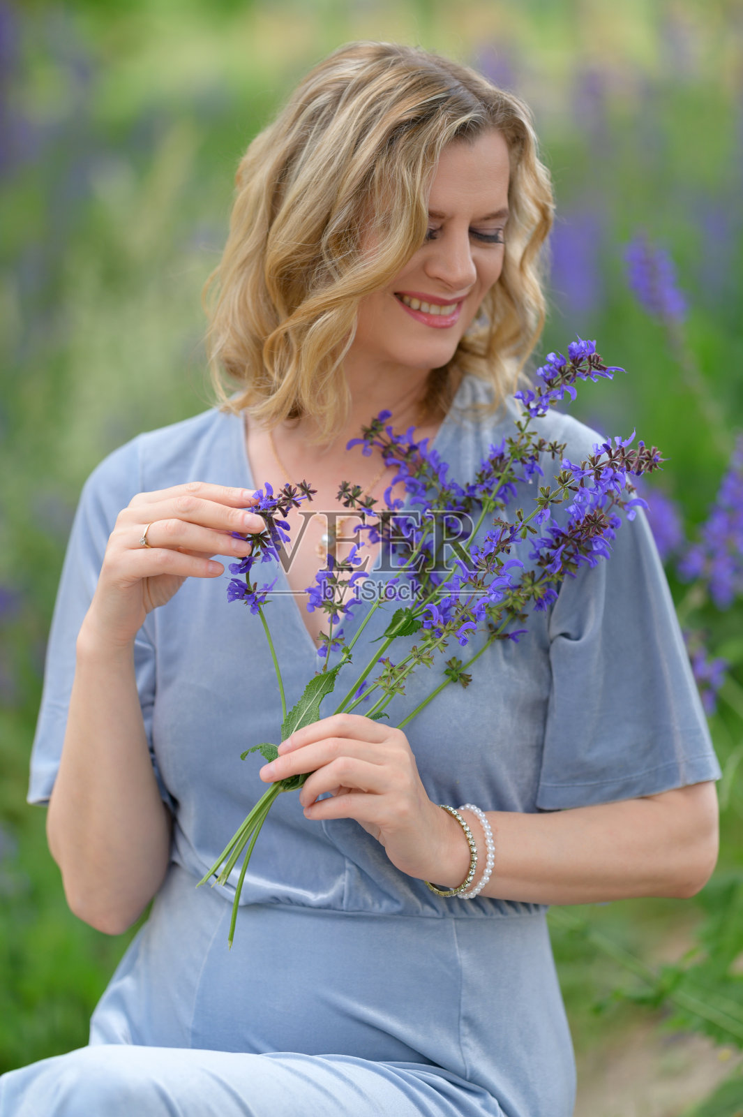 美丽的模特在春天或夏天坐在公园里。蓝色的裙子，紫色的花，稻草包，笔记本照片摄影图片