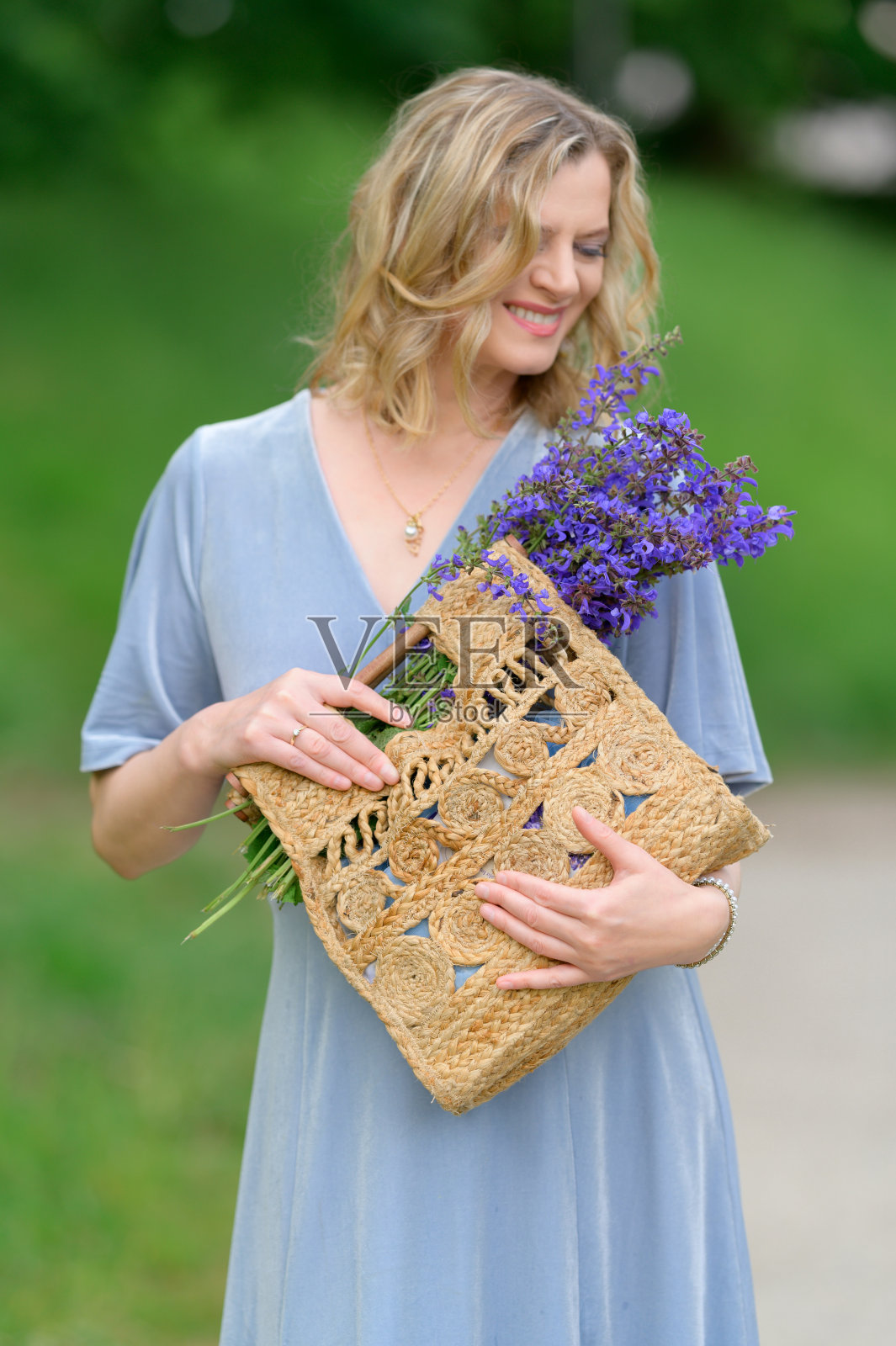 美丽的模特在春天或夏天在公园散步。蓝色的裙子，紫色的花，草编的袋子照片摄影图片