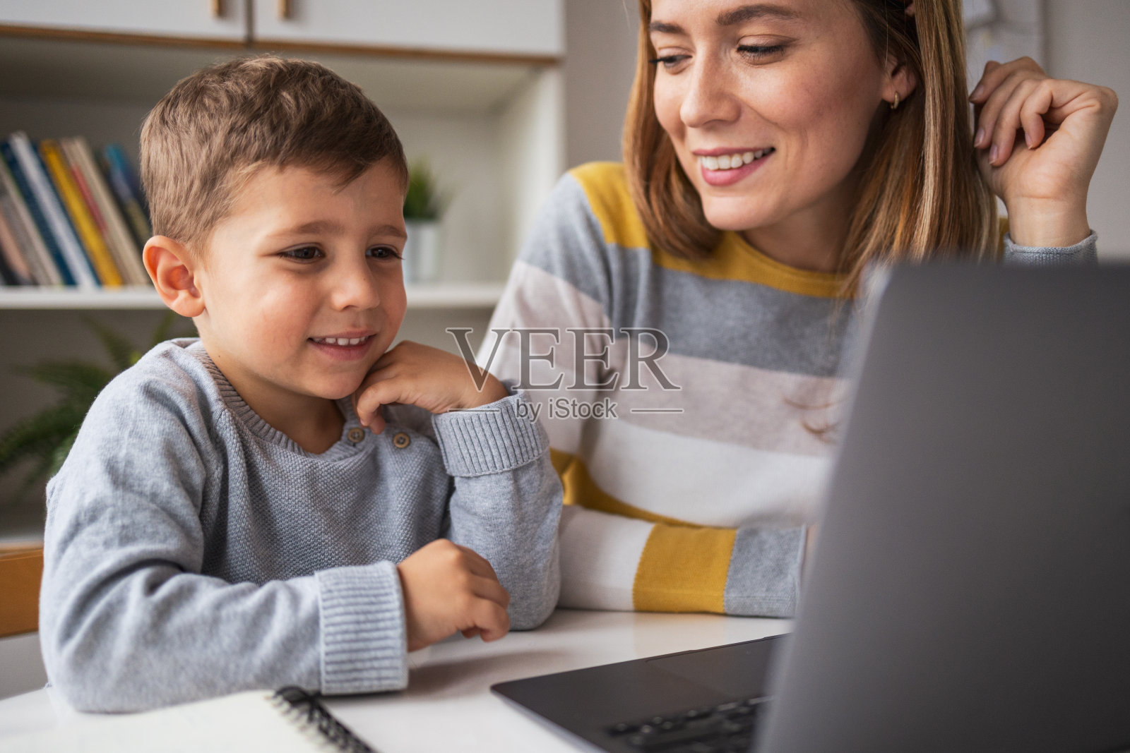 快乐的母亲看着儿子在公寓的桌子上使用笔记本电脑照片摄影图片