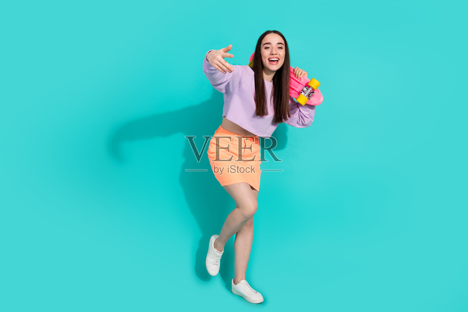 可爱的年轻女士手持滑板舞蹈的全身照片，穿着时尚的紫色服装，孤立于海蓝宝石色背景照片摄影图片