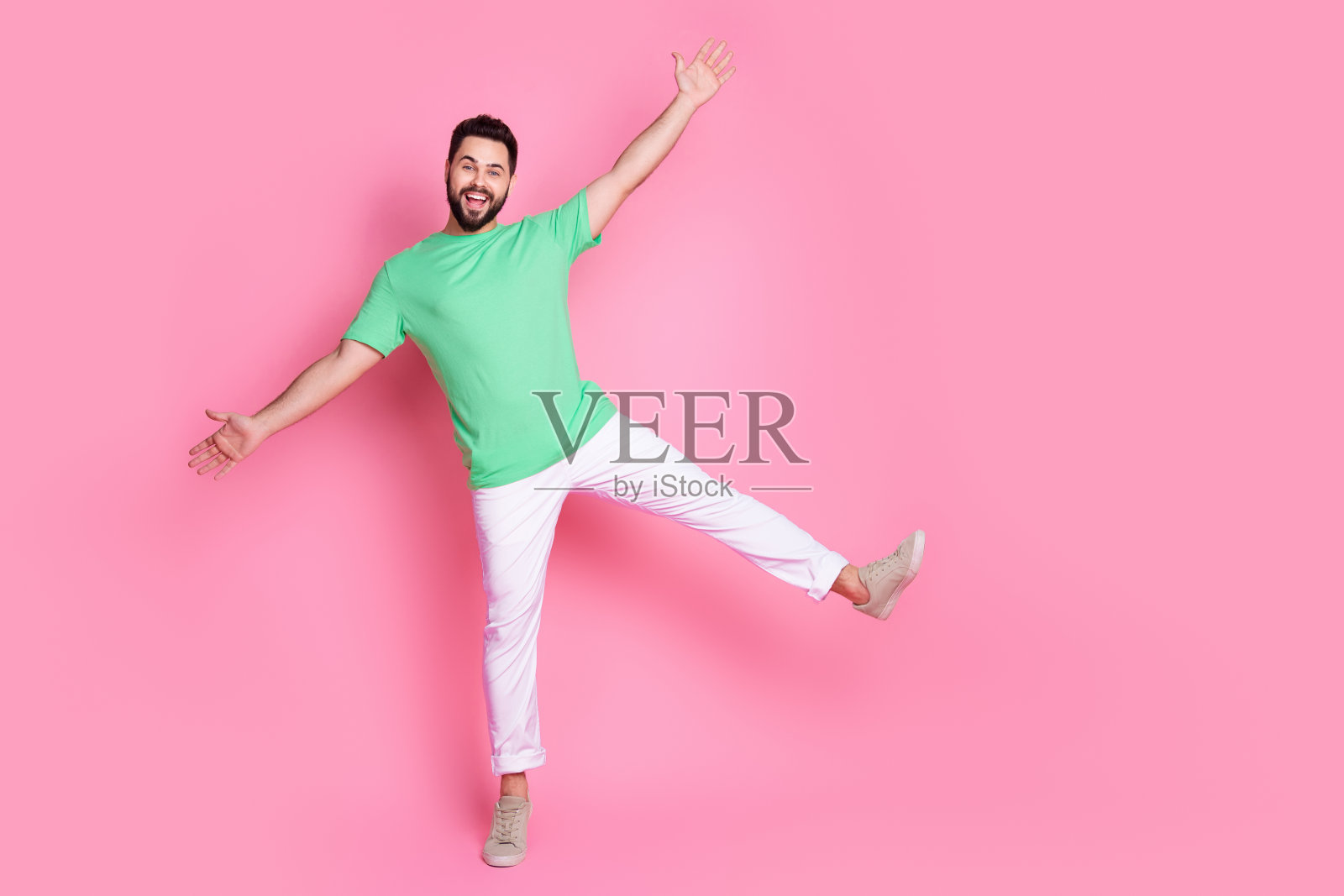全尺寸照片的好积极的家伙穿着绿色t恤白色裤子跳舞在一条腿上，双手在粉红色的背景孤立照片摄影图片