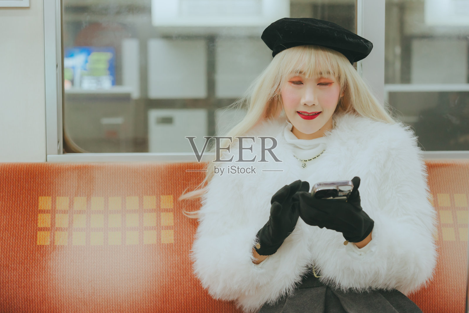 一个穿着白色皮大衣和黑色贝雷帽的女人坐在火车上看她的手机。照片摄影图片