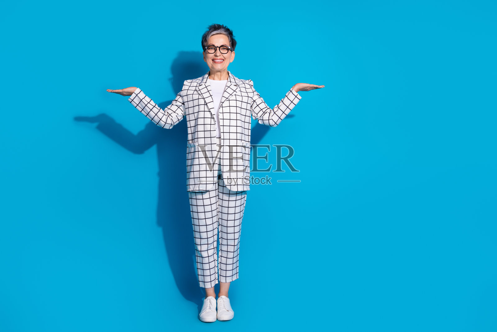 照片中，迷人的开朗女子穿着白色时髦的衣服，双臂抱着空旷的空间，孤立在蓝色的背景上照片摄影图片