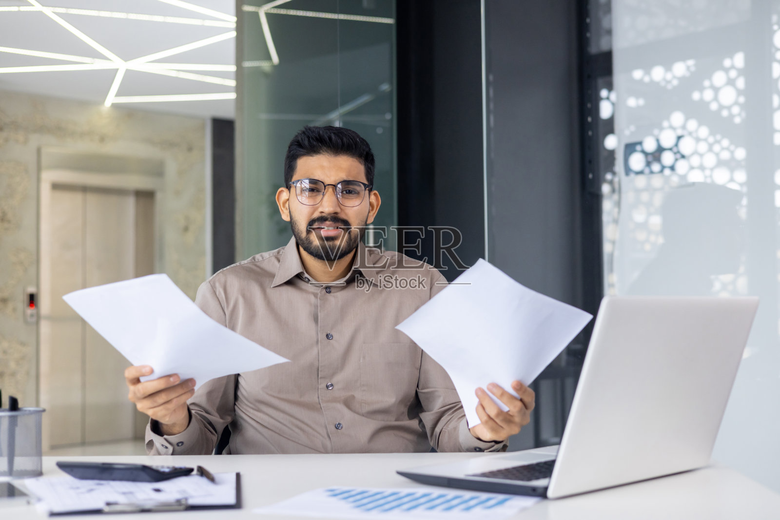 困惑的商人拿着文件在现代办公室的笔记本电脑前工作照片摄影图片