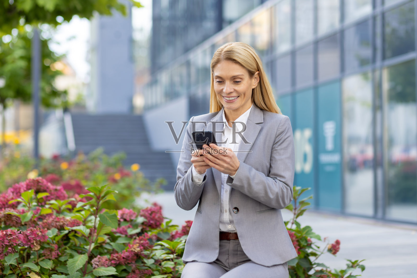 一位年轻漂亮的女商人坐在办公大楼附近的长椅上，微笑着使用手机照片摄影图片