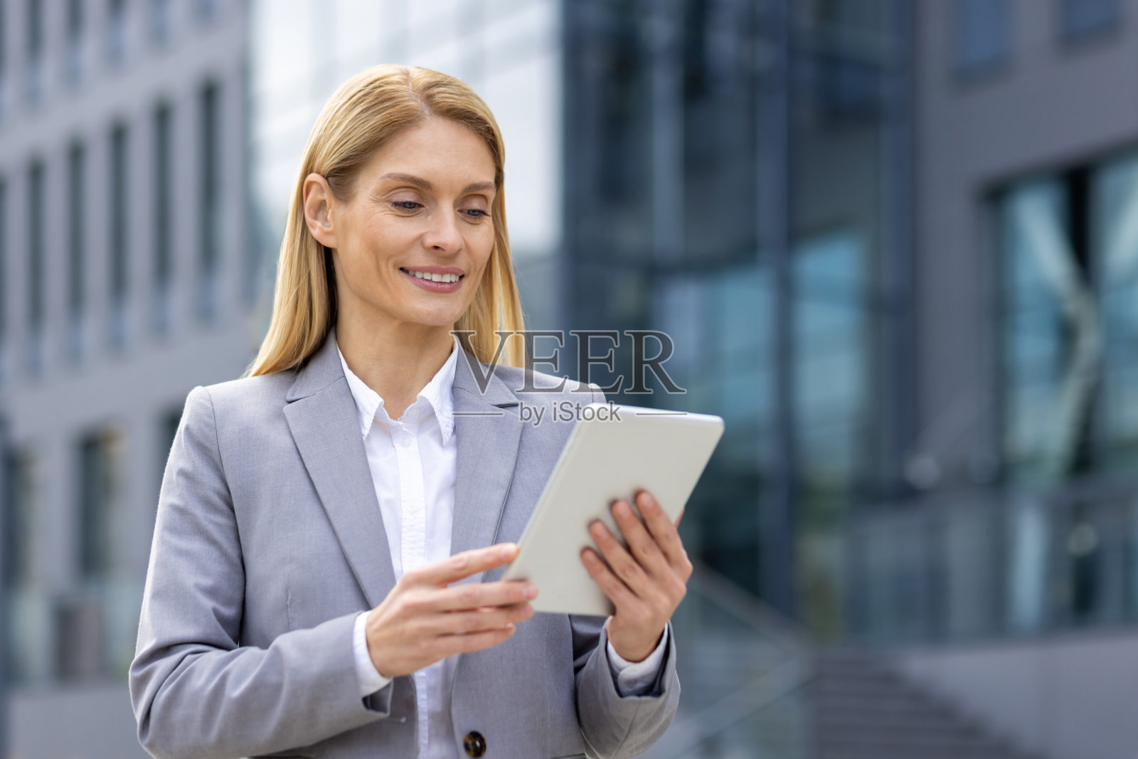 在城市里，一个自信的女商人站在户外，手里拿着一个平板电脑。她身穿灰色西装，在现代商业环境中散发出专业和成功的气息照片摄影图片